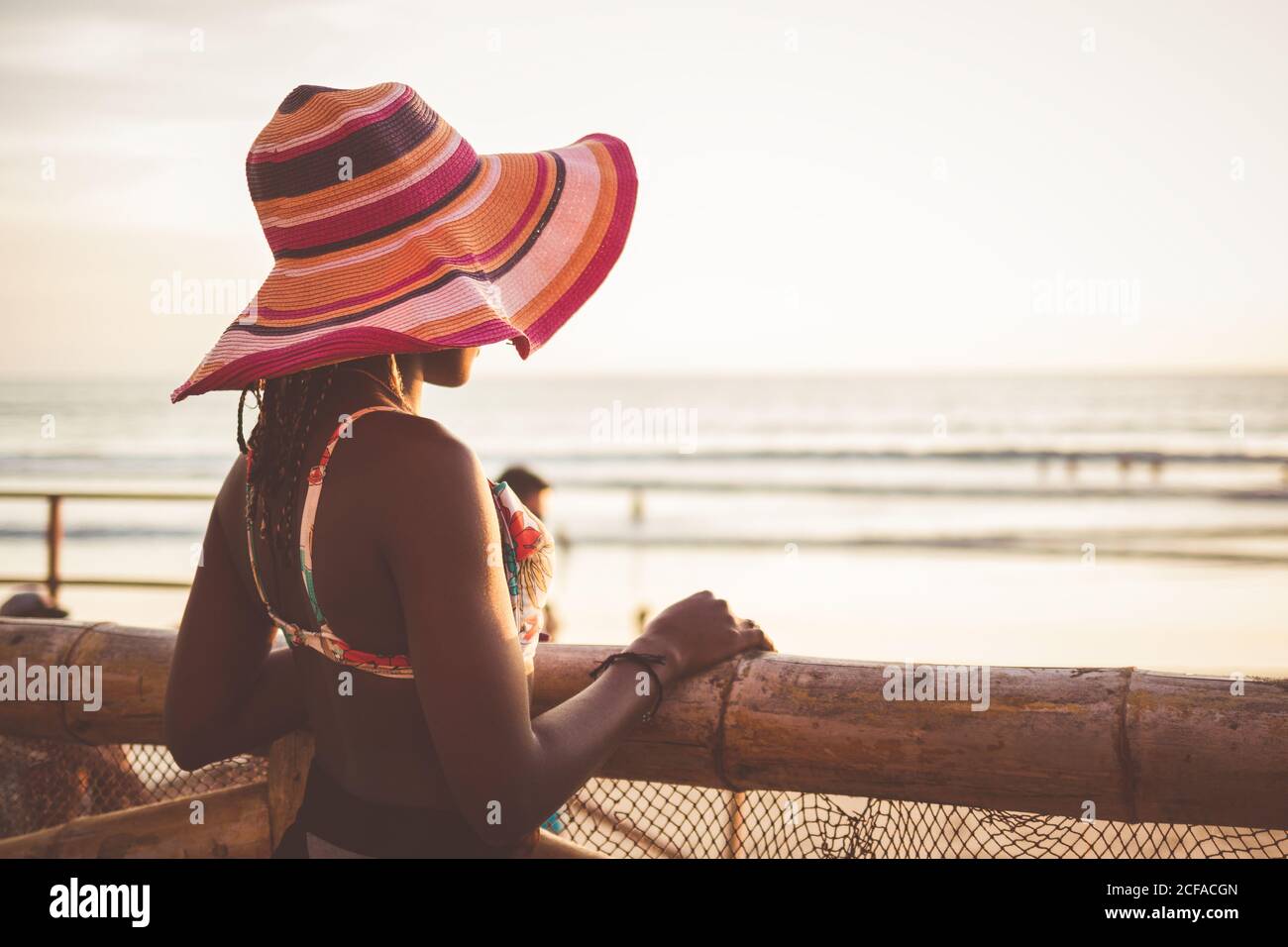 Seitenansicht der entspannten jungen afroamerikanischen Frau in bunt  Breiter gestreifter Hut mit Krempe und Bikini-Oberteil, das bei Holz steht  Zaun am Strand und genießen Seestück Stockfotografie - Alamy
