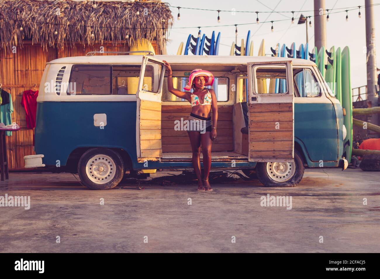 Junge afroamerikanische Besitzerin von lokalen Surfbrettverleih Tragen bunte Bikini-Top und Hut neben Vintage stehen Van mit Reihe von Surfbrettern im Hintergrund Stockfoto