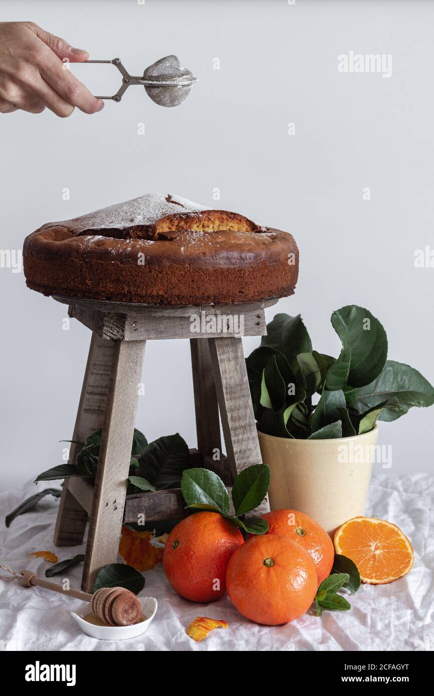 Crop anonyme Person Gießen Zucker Pulver mit Metall runden Tee Sieb über frischen appetitlichen Kuchen auf Holzhocker umgeben von Orange reife Mandarine und grüne Pflanze im Topf Stockfoto