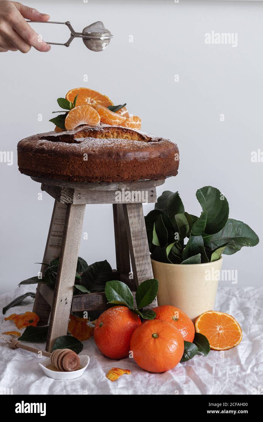Crop anonyme Person Gießen Zucker Pulver mit Metall runden Tee Sieb über frischen appetitlichen Kuchen auf Holzhocker umgeben von Orange reife Mandarine und grüne Pflanze im Topf Stockfoto