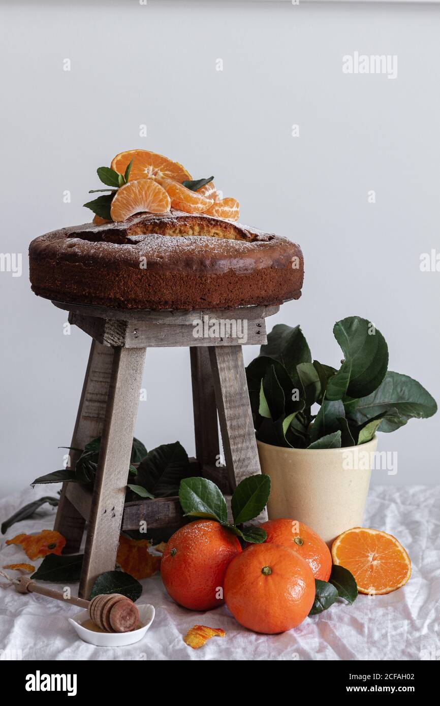 Malerisches Stillleben von frischem appetitlichen Schnitt und ganze Mandarine Frischer leckerer Kuchen auf kleinem Holzhocker und grüner Pflanze Im Topf Stockfoto
