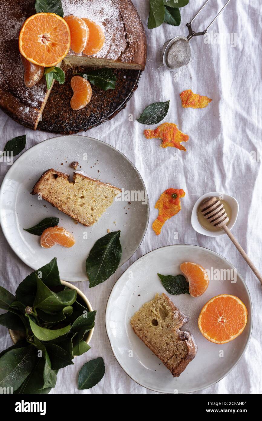 Von oben appetitlich süßer Kuchen und reife orange Mandarine geschnitten Und serviert auf weißen Tellern auf einem mit Pflanzen dekorierten Tisch Stockfoto