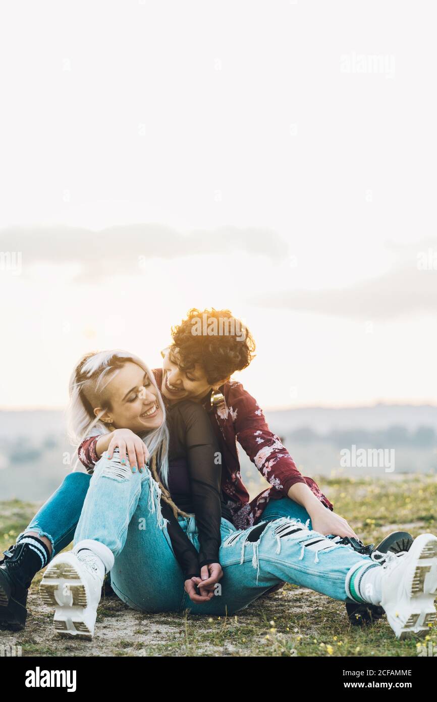 Lachend enge Freundinnen in stilvollen Casual Outfits sitzen auf grün Gras in Umarmung mit Himmel auf Hintergrund Stockfoto