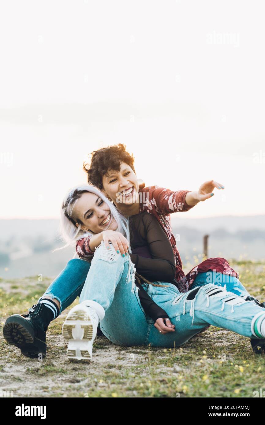Lachend enge Freundinnen in stilvollen Casual Outfits sitzen auf grün Gras in Umarmung mit Himmel auf Hintergrund Stockfoto