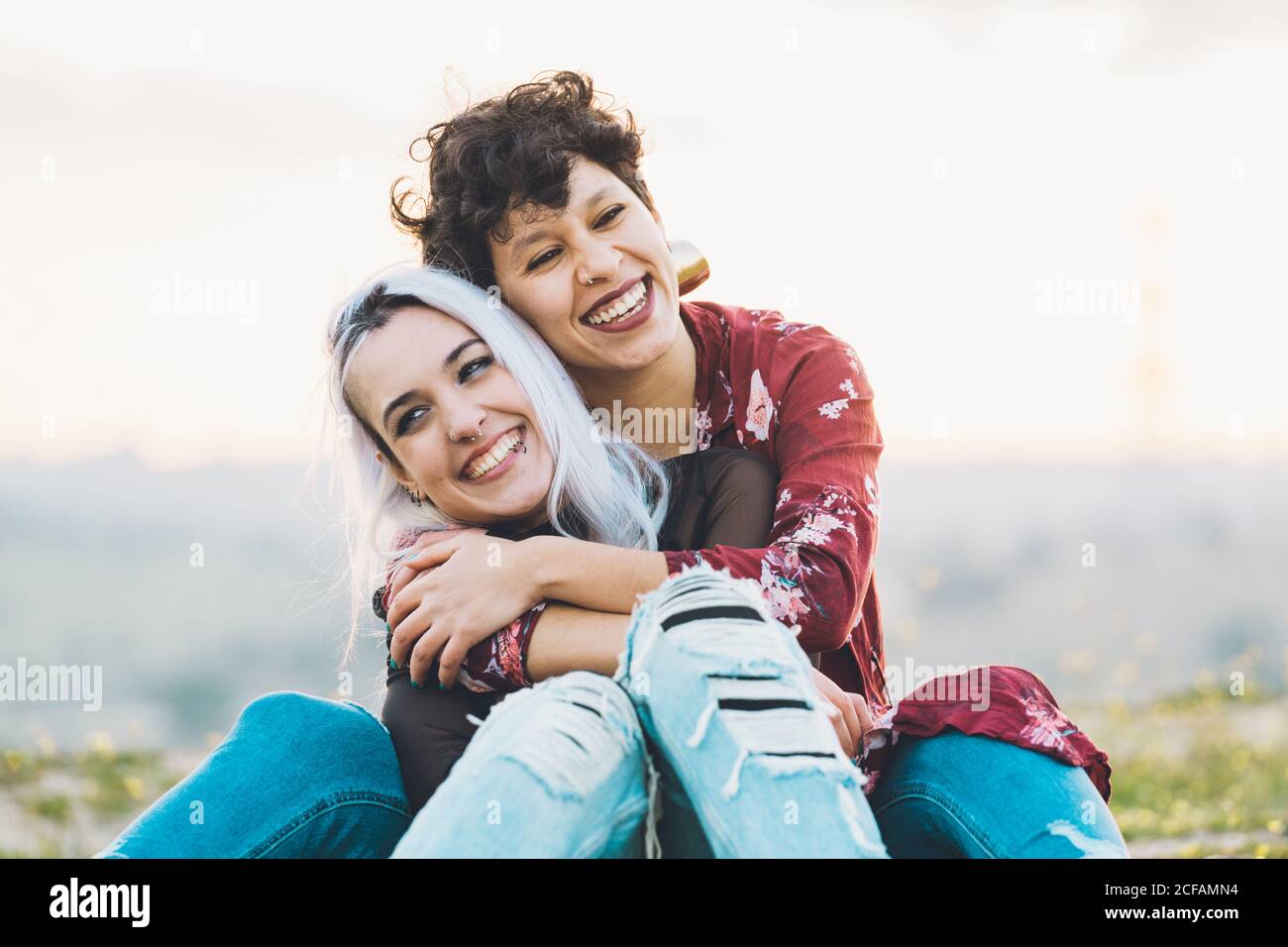 Lachend enge Freundinnen in stilvollen lässigen Outfits sitzen umarmt jeder Andere auf grünem Gras mit Himmel auf Hintergrund Stockfoto
