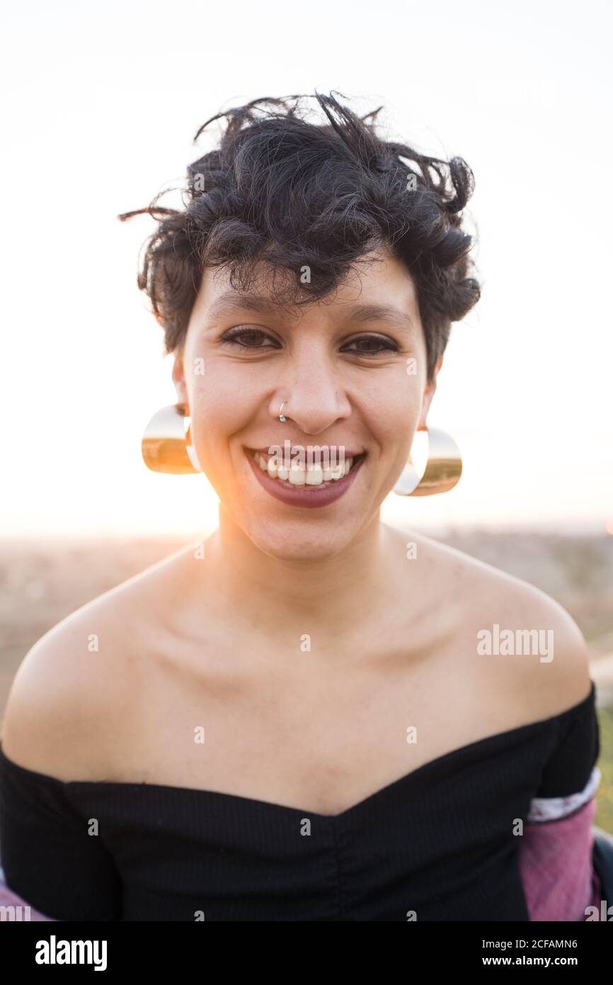 Schwarze kurzhaarige weibliche Teenager mit Ohrringen und piercing Lachen Und Blick auf die Kamera auf verschwommenem Hintergrund der Natur Stockfoto