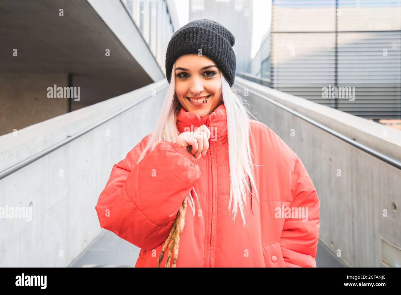 Blonde junge Hipster Frau in stilvollen roten warmen Jacke und Hut steht gegen moderne Gebäude Stockfoto