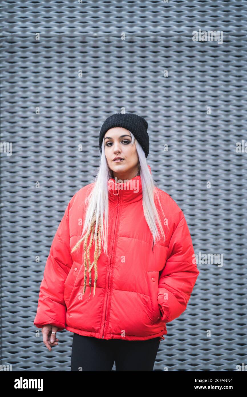 Blonde junge Hipster Frau in stilvollen roten warmen Jacke und Hut stehen vor modernen Metall Wand Hintergrund Stockfoto