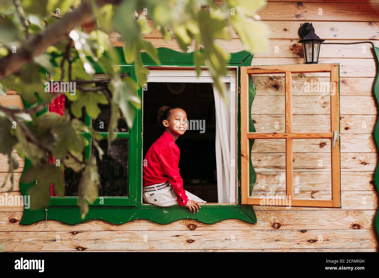 Fröhliches schwarzes Kind in rotem Hemd und weißer Hose suchen Durch das offene Fenster des ländlichen Holzhauses Stockfoto