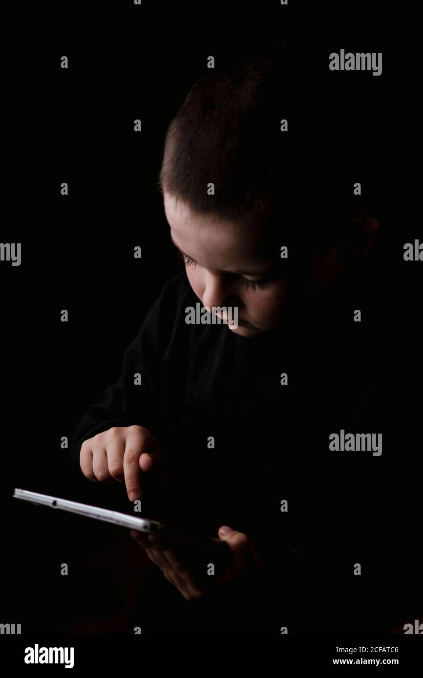 Liebenswert ernst Kind in Freizeitkleidung halten Tablet in den Händen Mit entschlossenem Blick isoliert auf schwarzem Hintergrund Stockfoto