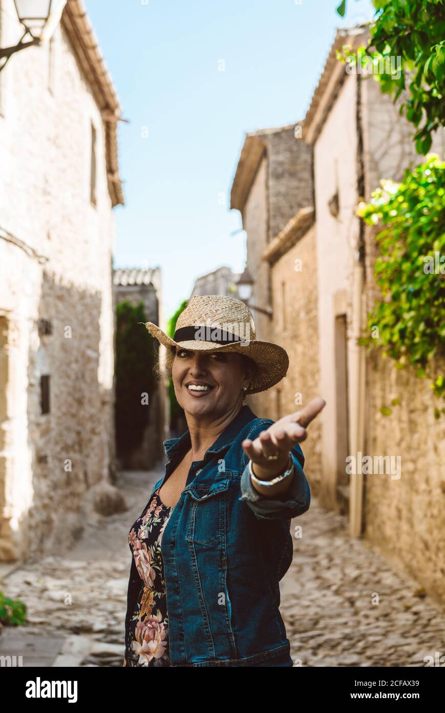 Frau trägt einen Hut und schaut auf die Kamera und reicht die Hand Zur Kamera, während in den Straßen eines mittelalterlichen Dorfes Mit Wandkletterer Werk in Europa Stockfoto