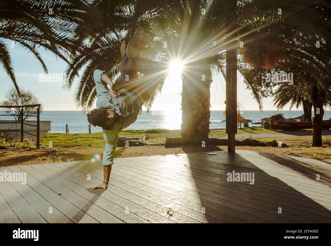 Moderne männliche Tänzerin in stilvoller Casual-Kleidung, die zur Hand steht Mit gekreuzten Beinen über Kopf auf der Terrasse des Hauses Mit tropischen Palmen am Meer und Sonnenstrahlen im Hintergrund Stockfoto