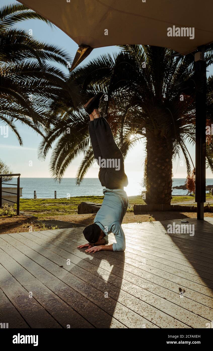 Moderne männliche Tänzerin in stilvoller Casual-Kleidung, die zur Hand steht Mit gekreuzten Beinen über Kopf auf der Terrasse des Hauses Mit tropischen Palmen am Meer und Sonnenstrahlen im Hintergrund Stockfoto