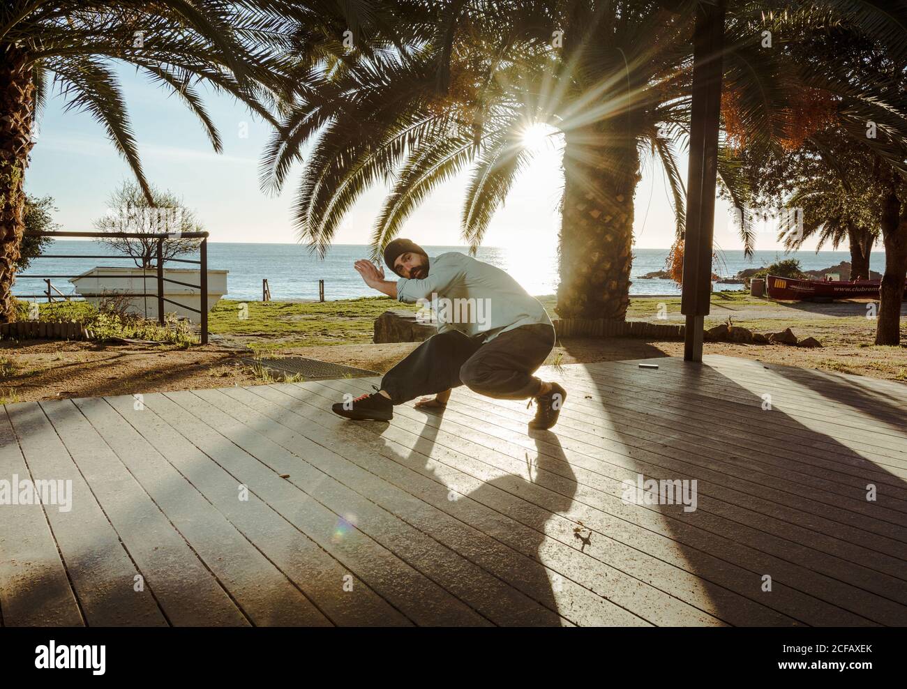 Moderne männliche Tänzerin in stilvoller Casual-Kleidung, die zur Hand steht Mit gekreuzten Beinen über Kopf auf der Terrasse des Hauses Mit tropischen Palmen am Meer und Sonnenstrahlen im Hintergrund Stockfoto
