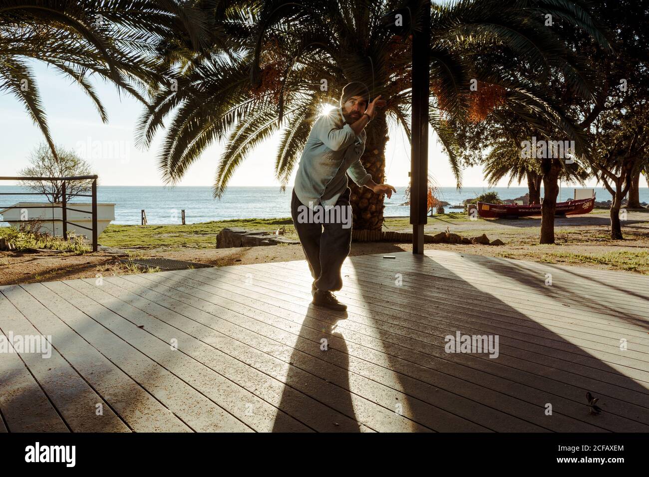 Moderne männliche Tänzerin in stilvoller Casual-Kleidung, die zur Hand steht Mit gekreuzten Beinen über Kopf auf der Terrasse des Hauses Mit tropischen Palmen am Meer und Sonnenstrahlen im Hintergrund Stockfoto