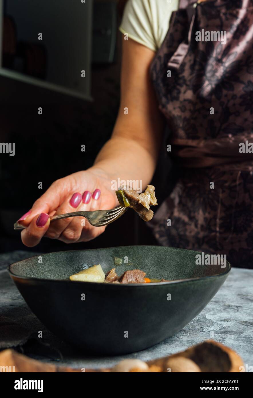 Crop weibliche kochen in Schürze mit Scheibe Rindfleisch auf Gabel steht neben dem Tisch mit Schüssel mit köstlichen Rindfleisch Eintopf In der Küche Stockfoto