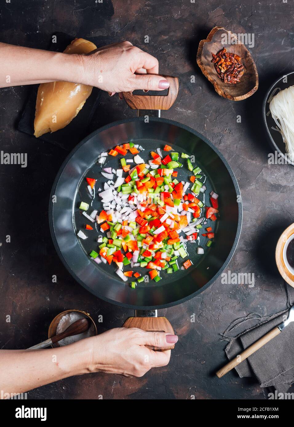 Draufsicht der Ernte anonyme Hausfrau schütteln Pfanne und Mischen Gehackte Paprika und Zwiebel beim Kochen Nudeln mit Huhn für Abendessen Stockfoto