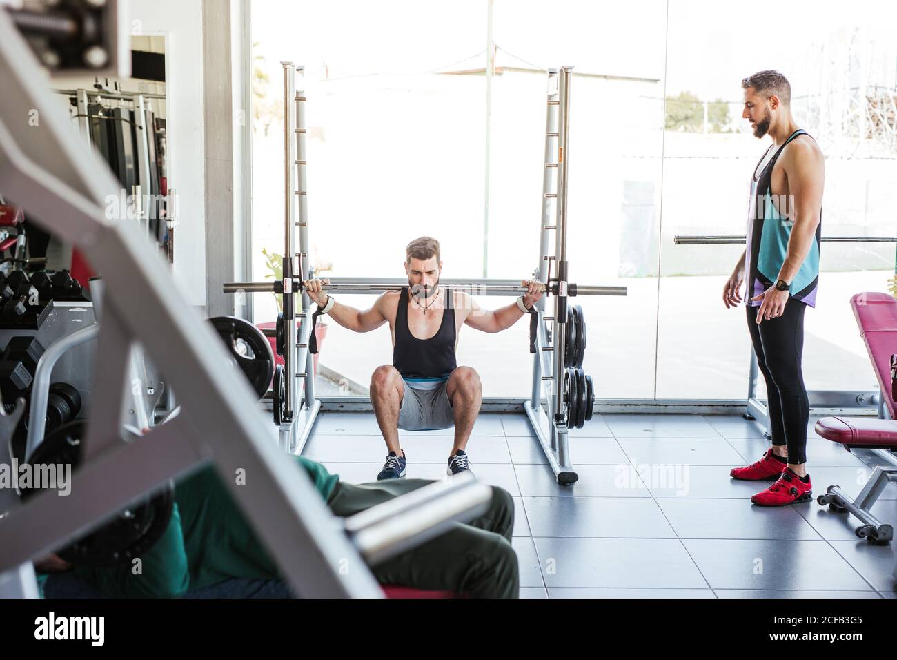 Kraftvoller entschlossener Sportler, der mit Langhantel an der Hocke trainiert Beim Gewichtheben mit Personal Coach im modernen Fitnessstudio Stockfoto