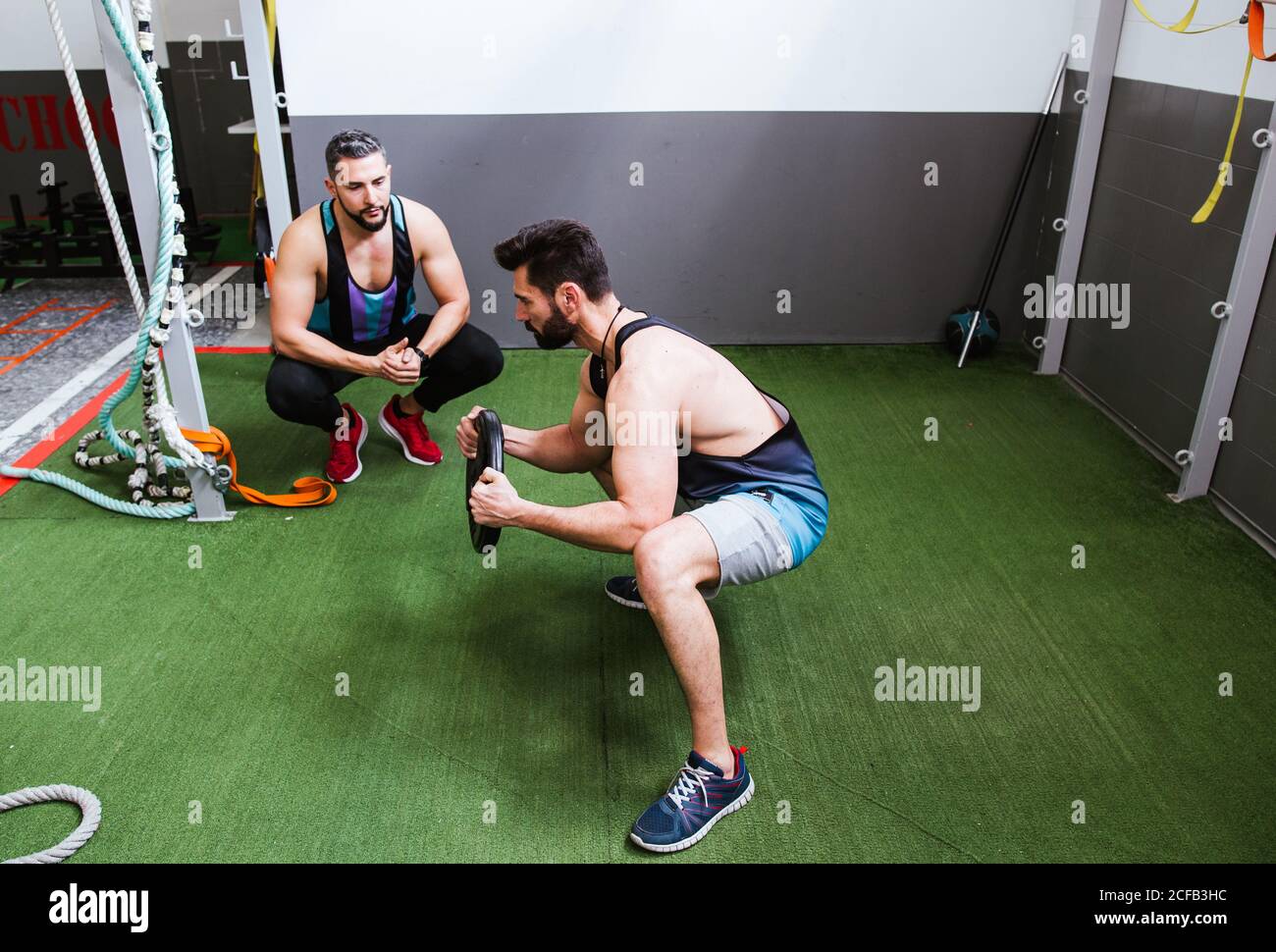 High-Angle-Seitenansicht des fokussierten starken Sportlers beim Hocken Übung mit Gewichtscheibe beim Training im Fitnessstudio mit Trainer Stockfoto