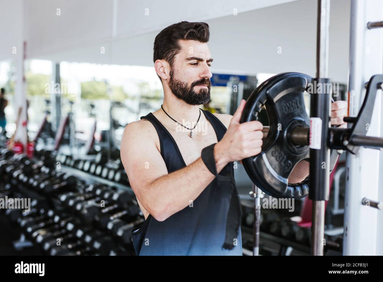 Muskulöser bärtiger Mann in Sportkleidung, der in der Nähe von Hockregal und steht Setzen Gewicht Scheibe auf Langhantel während der Vorbereitung für Gewichtheben Training Im modernen Fitnessraum Stockfoto