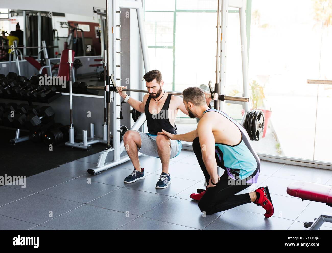 Kraftvoller entschlossener Sportler, der mit Langhantel an der Hocke trainiert Beim Gewichtheben mit Personal Coach im modernen Fitnessstudio Stockfoto