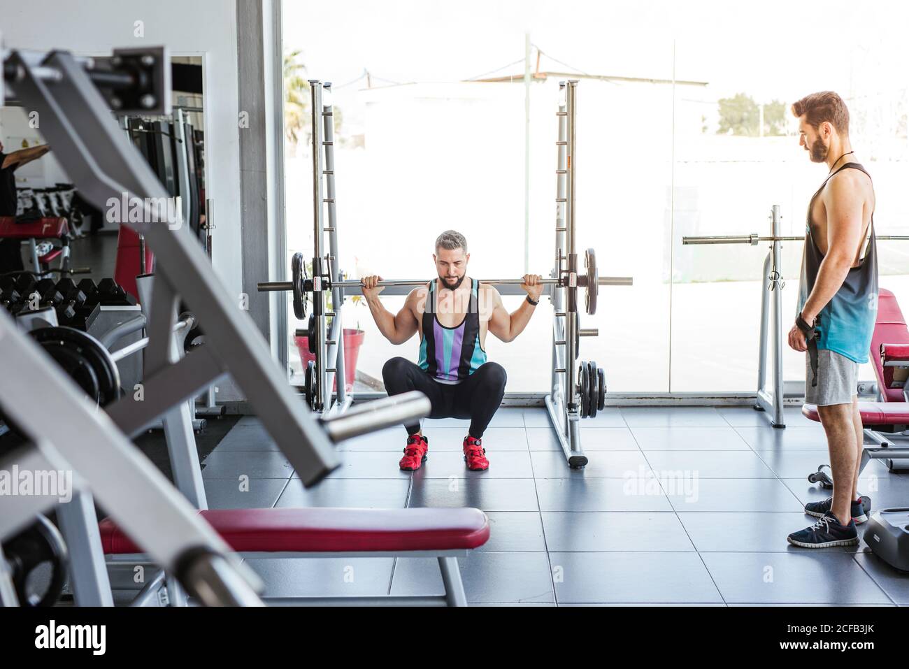 Kraftvoller entschlossener Sportler, der mit Langhantel an der Hocke trainiert Beim Gewichtheben mit Personal Coach im modernen Fitnessstudio Stockfoto