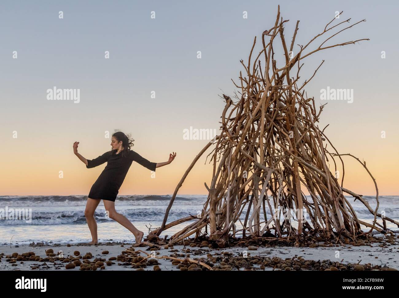 Junge Tänzerin an der Sandküste mit Holzzweigen für platziert Lagerfeuer in der Nähe von wehenden Meer und blauem Himmel Stockfoto