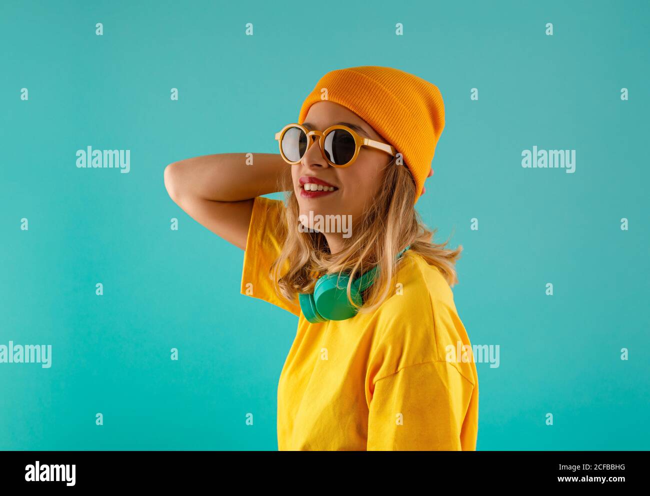 Seitenansicht der glücklichen jungen niedlichen weiblichen in gelben Outfit Und orange Beanie wegschauen mit Sonnenbrille Sonnenbrillen gegen bunte Türkisfarbener Hintergrund Stockfoto