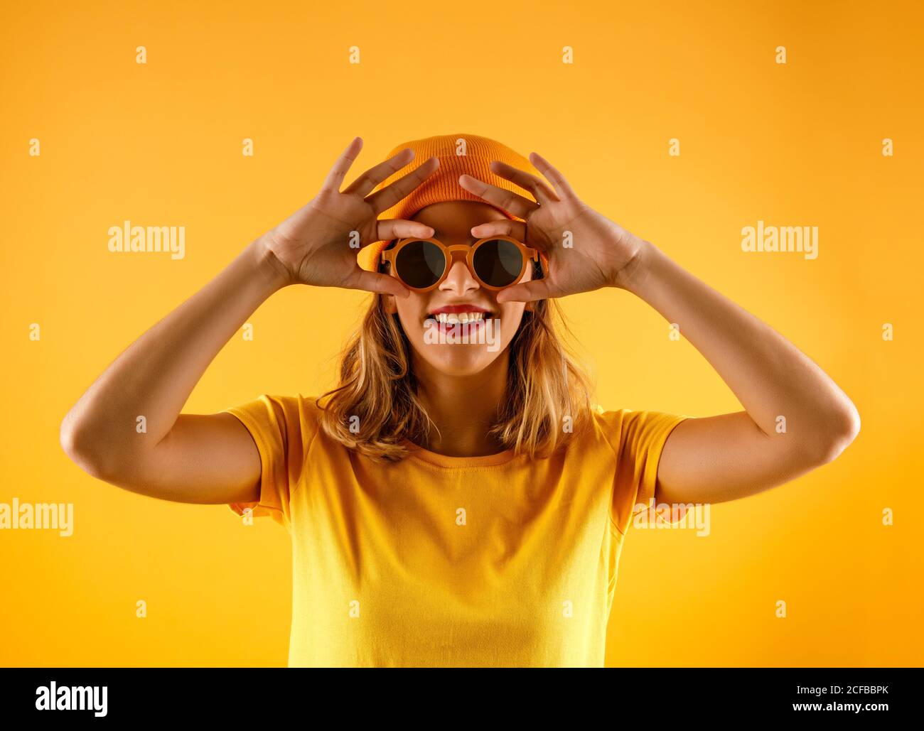 Fröhliche junge Frau in heller Kleidung und stylischer Sonnenbrille lächelt und blickt vor orangefarbenem Hintergrund auf die Kamera Stockfoto