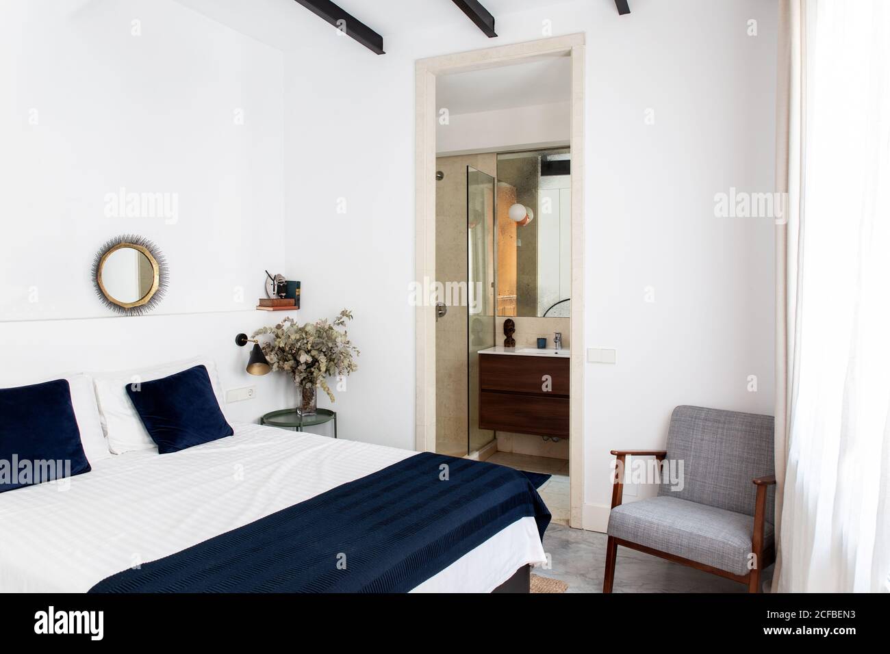 Komfortables Bett mit weißer und blauer Bettdecke und weichem Sessel Befindet sich in der Nähe von Bad Tür in gemütlichen Schlafzimmer der modernen Wohnung Stockfoto