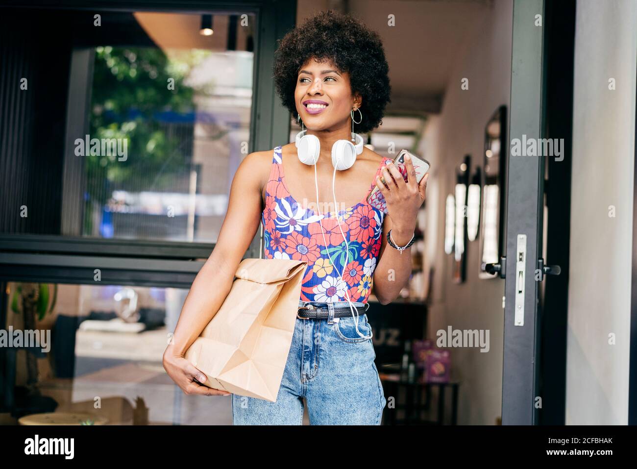 Ziemlich moderne afroamerikanische Frau mit Kopfhörern am Hals und Smartphone in den Händen halten Papiertüte mit Lebensmitteln zu gehen Beim Verlassen des Coffee Shops Stockfoto