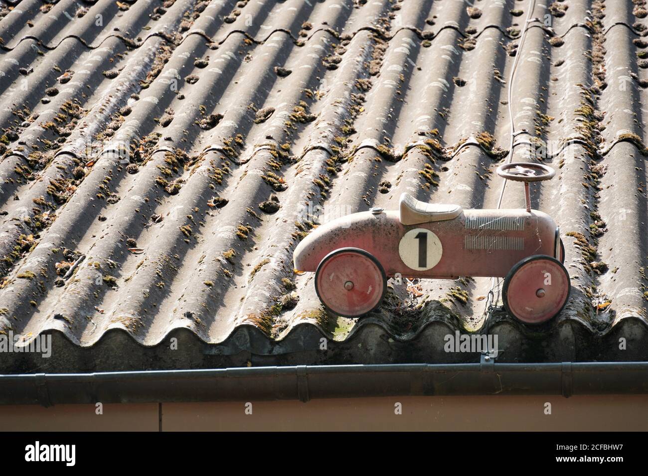 Ein rotes Schaukelauto, das auf einem alten grauen Korrugat-Faserzementdach mit Moos aufgestellt wurde. Das Auto ist altmodisch und hat die Nummer 1 bemalt. Stockfoto