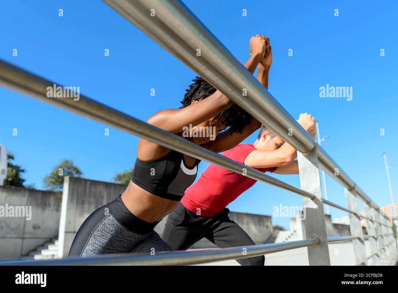 Von unten sehen Sie eine fröhliche afroamerikanische Fitnessfrau und einen lächelnden, sportlichen Mann, der neben einem Metallzaun steht und sich die Beine streckt, während Sie an einem sonnigen Tag mit blauem Himmel im Hintergrund gemeinsam trainieren Stockfoto