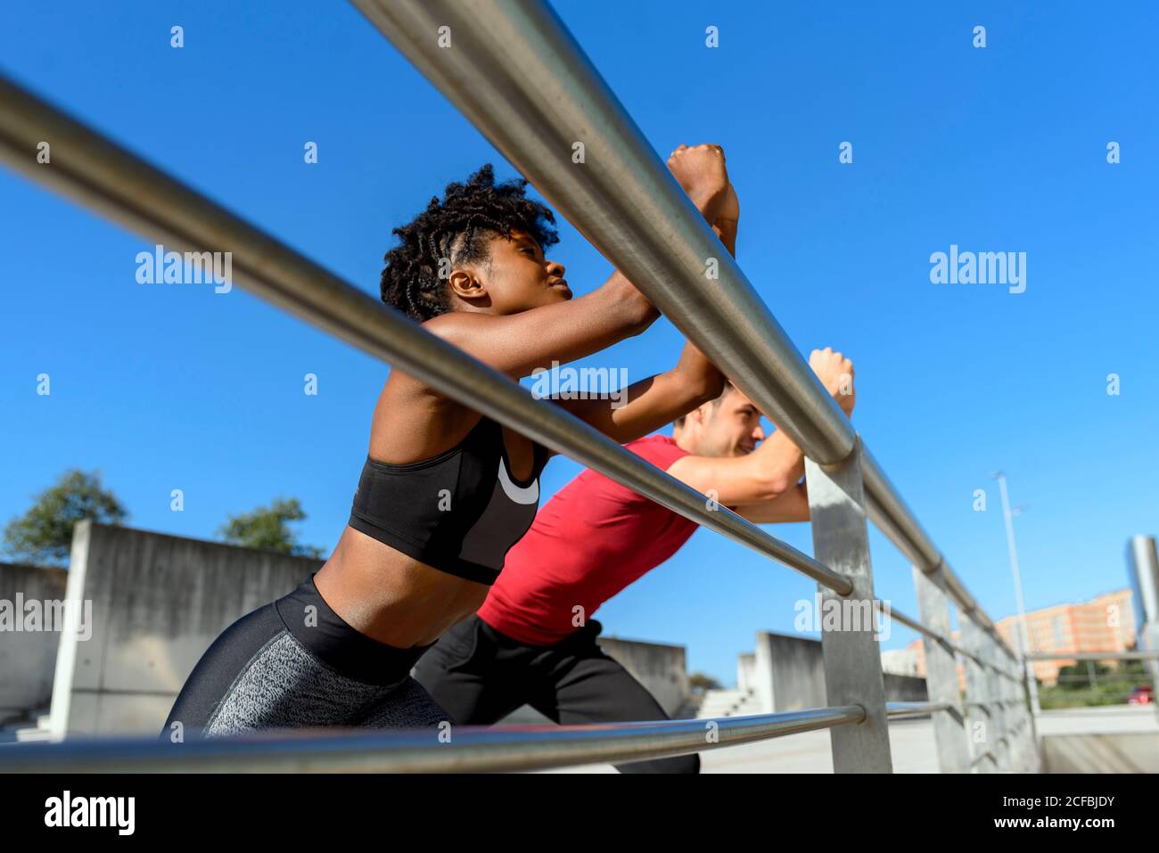 Von unten sehen Sie eine fröhliche afroamerikanische Fitnessfrau und einen lächelnden, sportlichen Mann, der neben einem Metallzaun steht und sich die Beine streckt, während Sie an einem sonnigen Tag mit blauem Himmel im Hintergrund gemeinsam trainieren Stockfoto