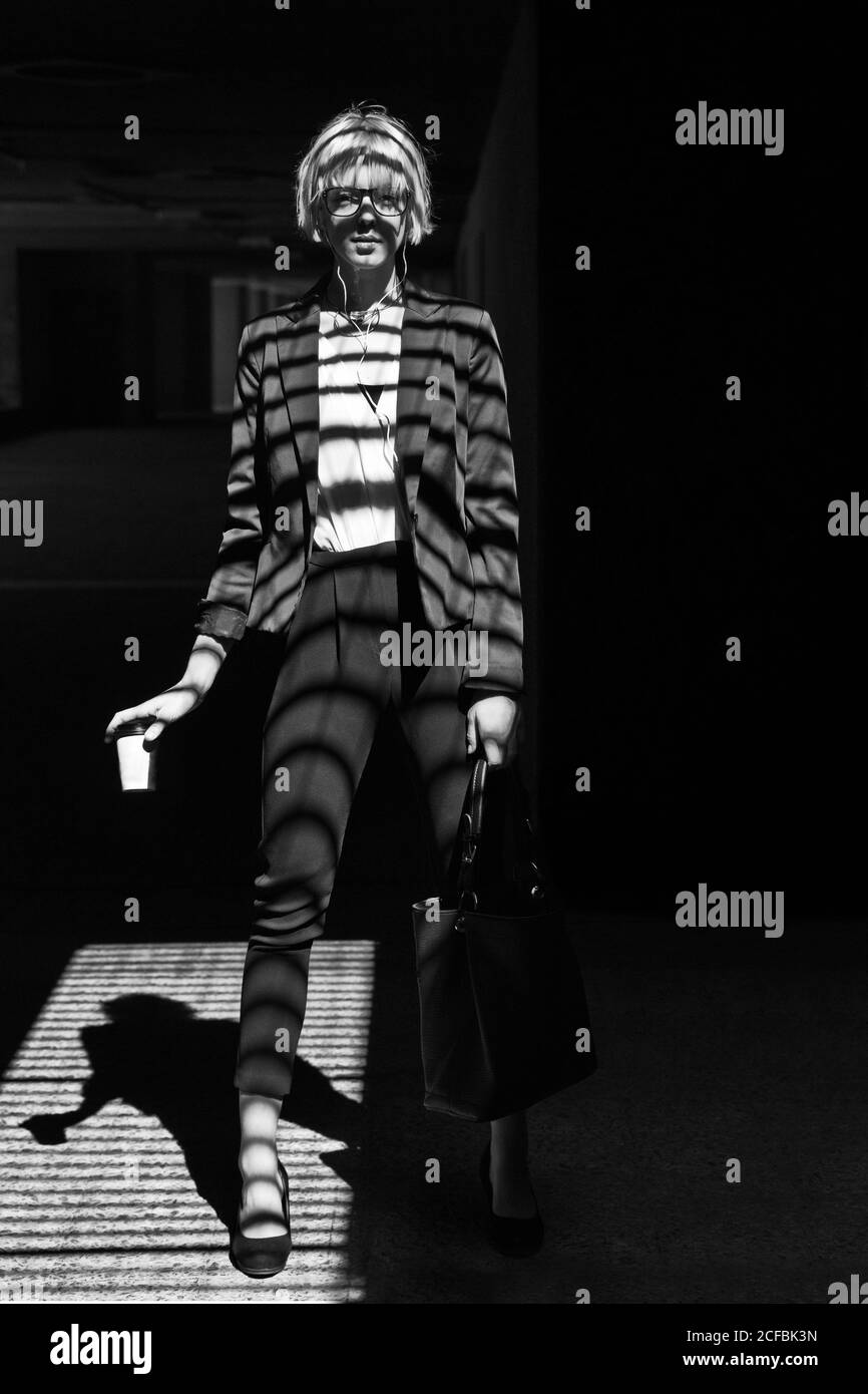 Schwarz-Weiß-Aufnahme einer stilvollen Frau in formeller Kleidung, die mit einem Papierbecher am Fenster steht und die Kamera anschaut Stockfoto