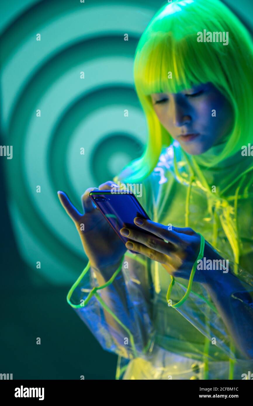 Junge asiatische Frau in futuristischen tragen und grüne Perücke mit Smartphone in fluoreszierendem Licht Stockfoto