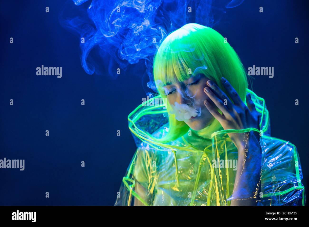 Junge ziemlich ungewöhnliche asiatische Frau in Kunststoff transparent Regenmantel und gelbes Haar Rauchen in fluoreszierendem Licht Stockfoto