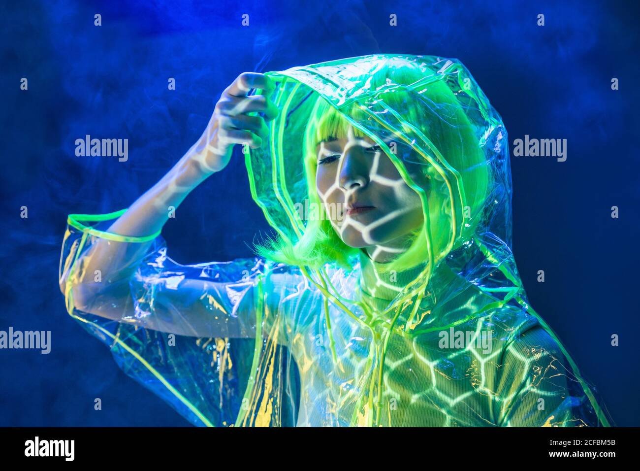 Junge ziemlich ungewöhnliche asiatische Frau in Kunststoff transparent Regenmantel und gelbes Haar Rauchen in fluoreszierendem Licht Stockfoto