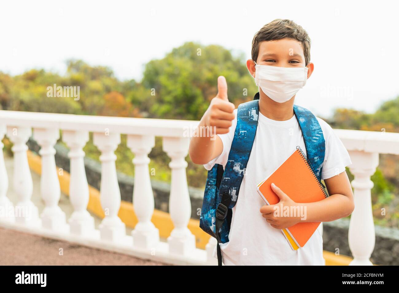 Portrait der Schule Kind trägt Schutzmaske für Verschmutzung oder Virus und zeigt Daumen nach oben, während auf den Schulbus am Morgen warten. Coron Stockfoto