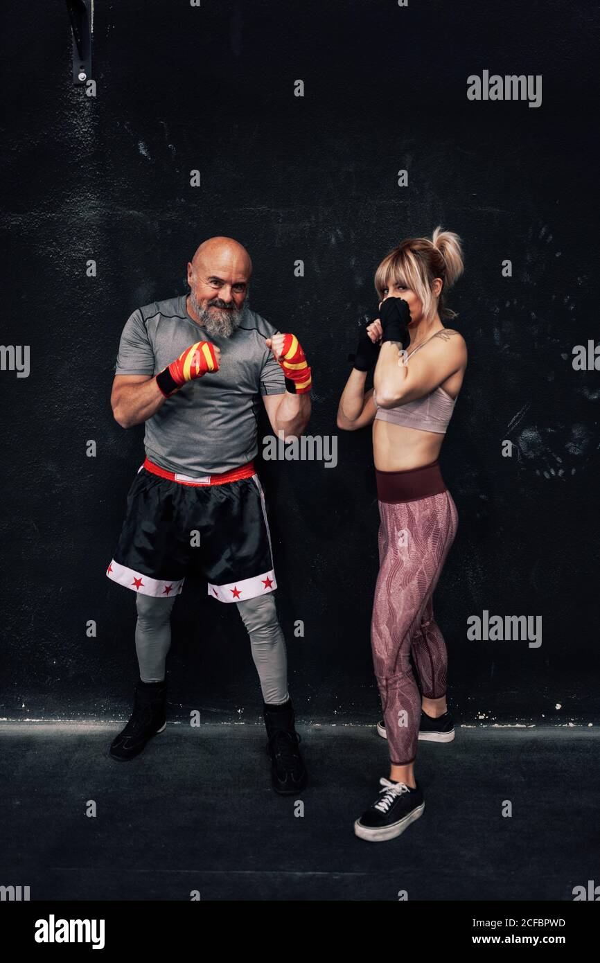 Positive starke männliche und weibliche Athleten mit eingehüllten Händen Bandagen stehen in Boxstellung und schauen gegen die Kamera Dunkler Hintergrund Stockfoto