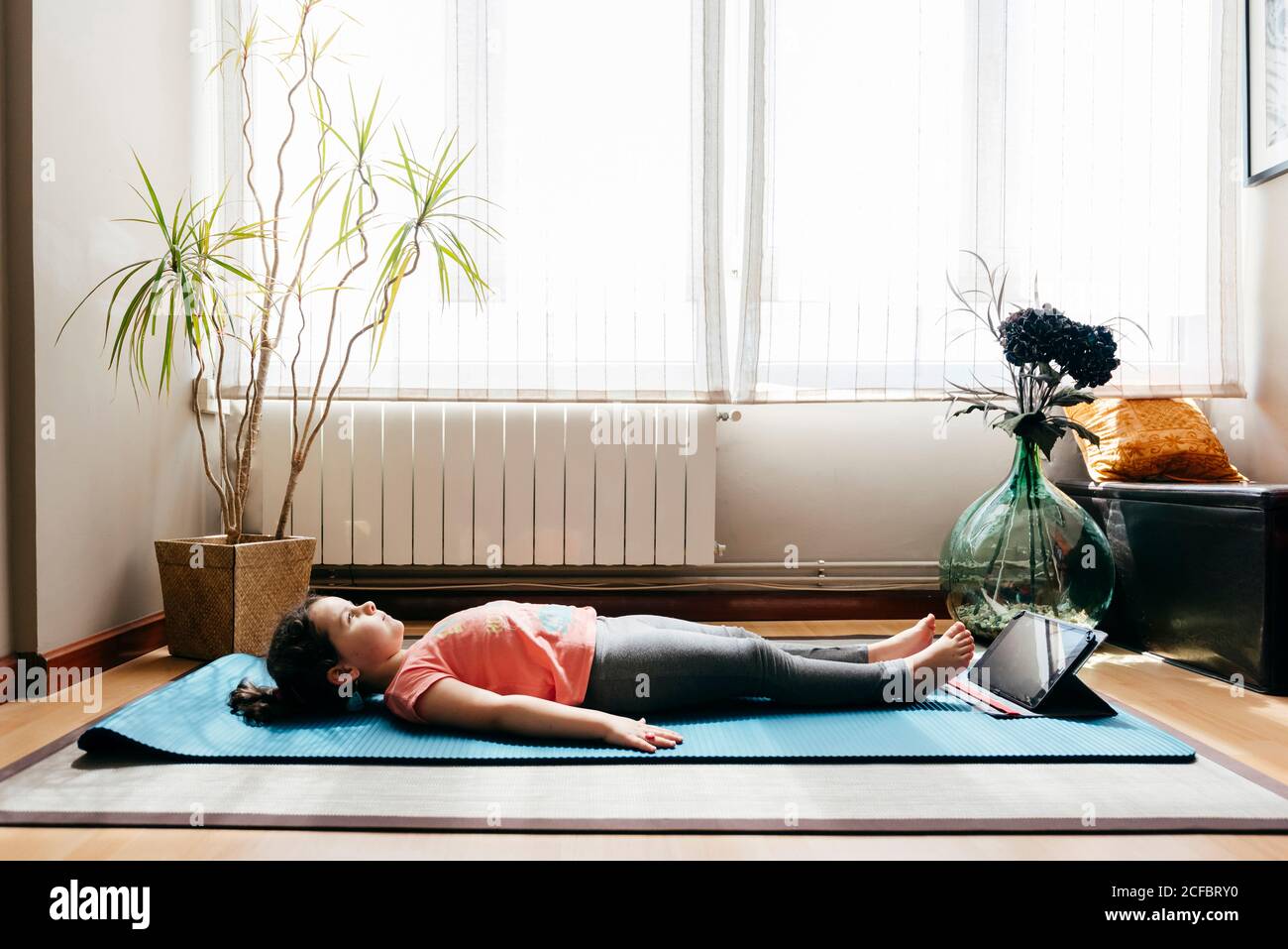 Seitenansicht des ruhigen kleinen Mädchens in Sportkleidung liegend Leiche Pose und entspannen beim Yoga in der Nähe Laptop zu üben Zu Hause Stockfoto