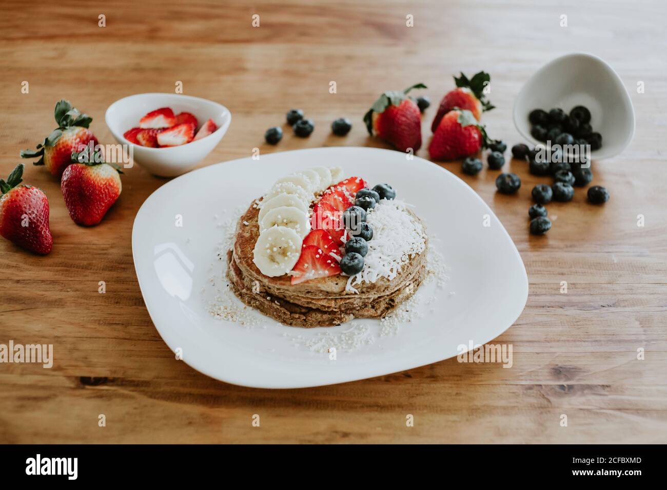 Stapel von leckeren Pfannkuchen serviert auf dem Teller mit Stücken Banane und Erdbeere und frische Heidelbeeren mit Kokosflocken während Frühstück Stockfoto