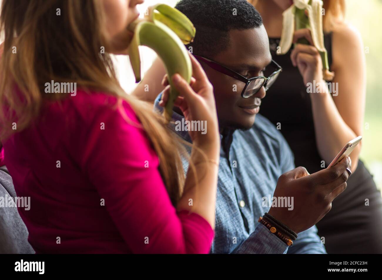 Dreiecksbeziehung. Zwei Frauen schauen auf schwarzen Mann und essen Banane, während Männer zwischen ihnen sitzen und Telefon Stockfoto