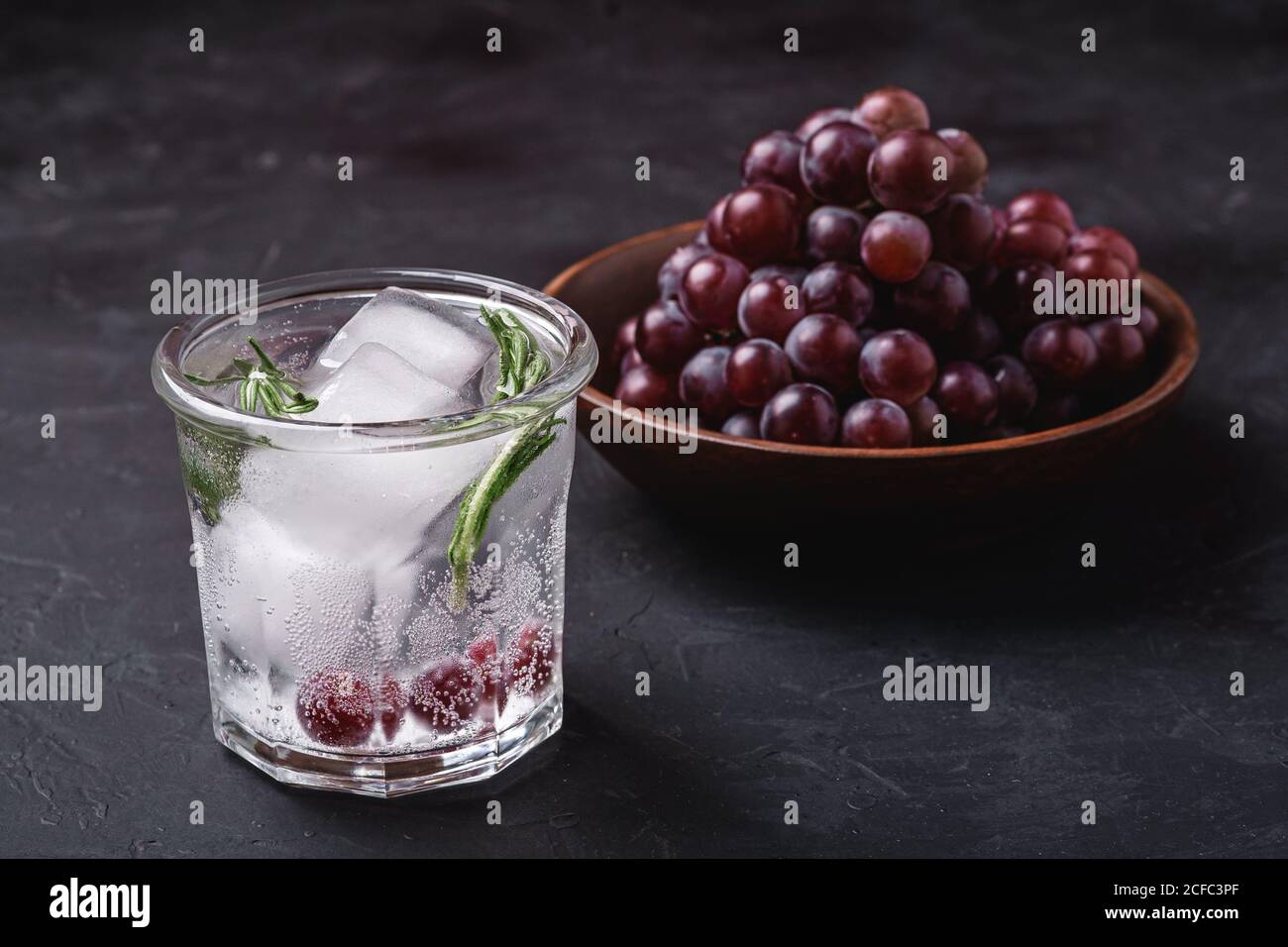 Frisches, eiskaltes kohlensäurehaltiges Wasser im Glas mit Rosmarinblatt in der Nähe einer Holzschale mit Traubenbeeren, dunkler Steinhintergrund, Winkelansicht Stockfoto