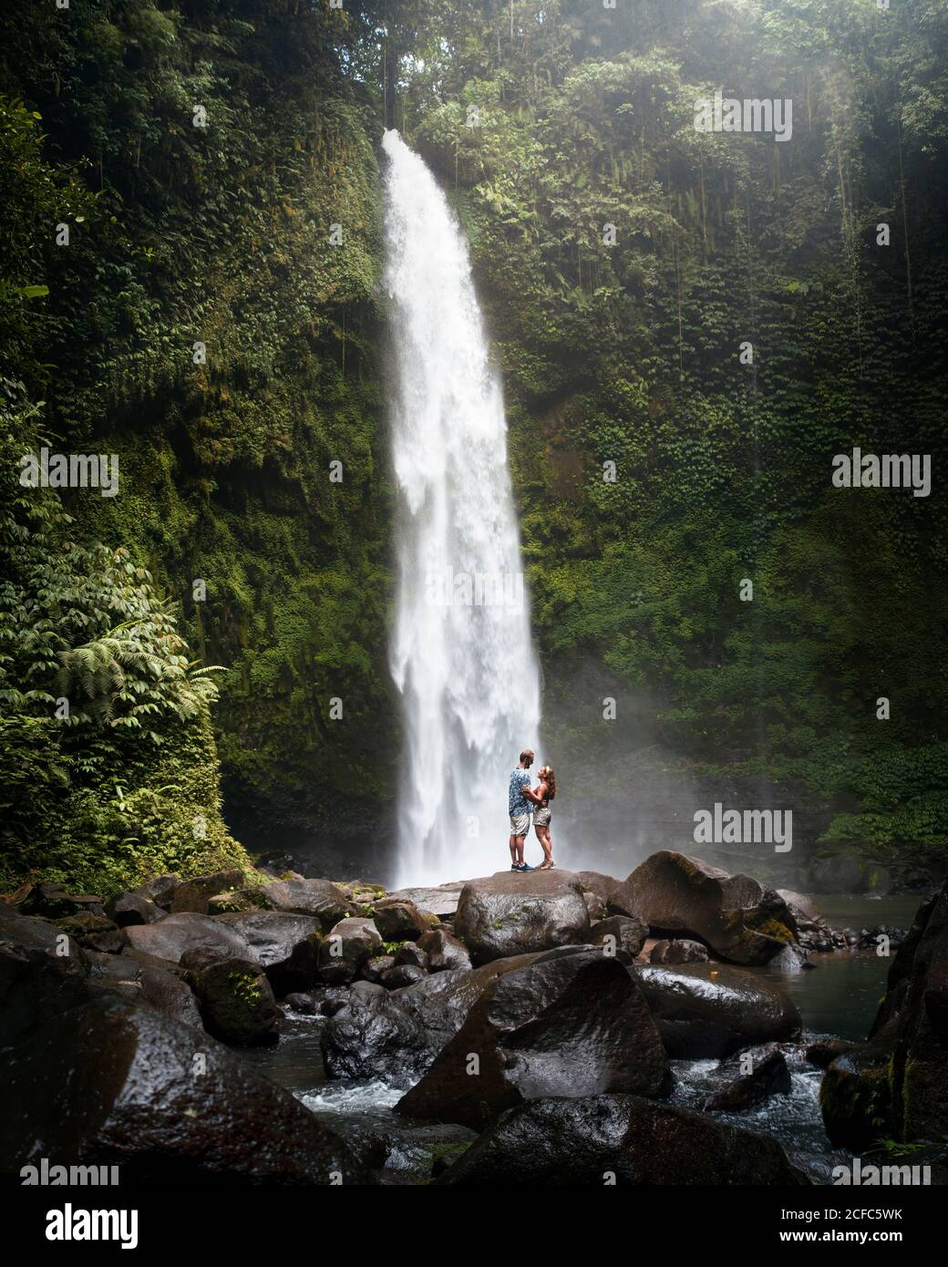 Seitenansicht von zwei Reisenden in legerer Kleidung, die darin stehen Vorne von einander auf rauem Felsen in der Nähe mächtig hoch Wasserfall zwischen grünen tropischen Pflanzen in Indonesien Stockfoto
