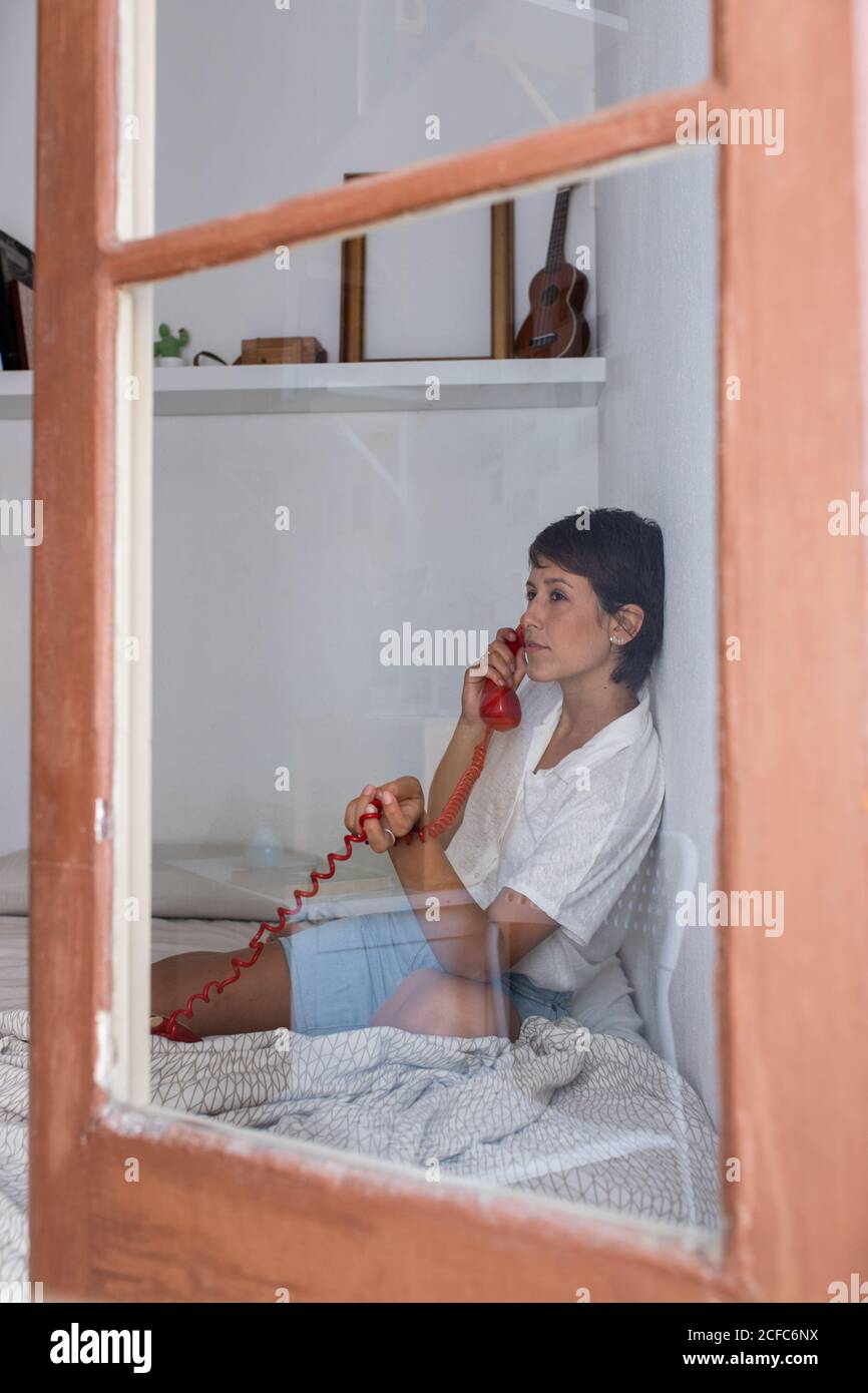 Junge Frau und nimmt Telefonanruf von vintage roten Telefon Während Sie auf dem Bett im gemütlichen Schlafzimmer durch Glas sitzen Stockfoto