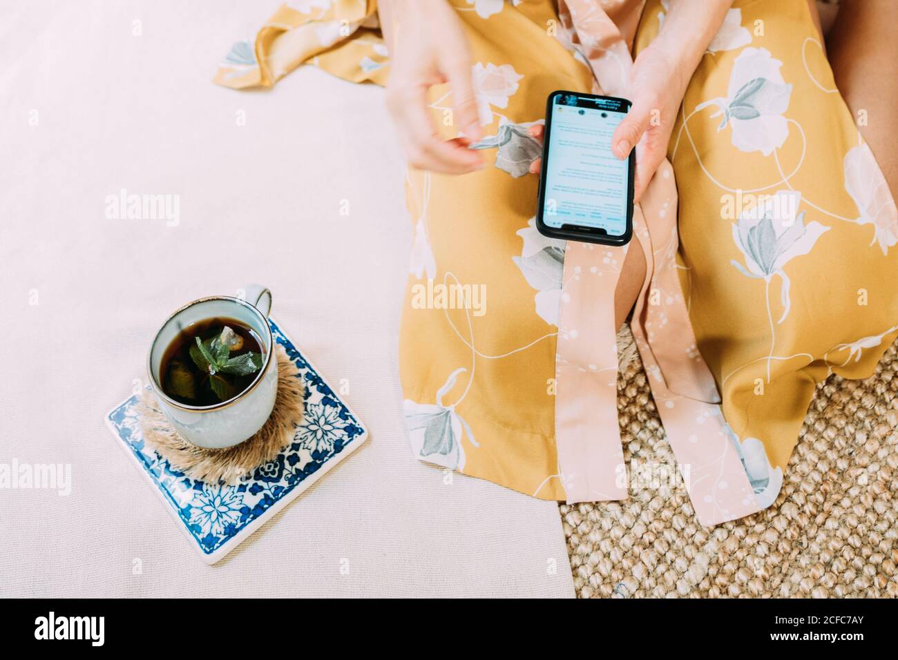 Von oben auf nicht erkennbare weibliche Surfen Handy während des Sitzens Auf Korbmatte in der Nähe Tasse heißen leckeren Tee mit Minze am Wochenende zu Hause Stockfoto