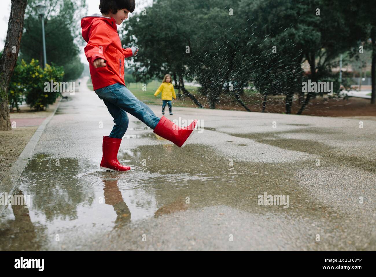 Liebenswert fröhliche Kinder in rot und gelb Regenmantel und Gummi Stiefel mit Spaß spielen in Pfütze in der Straße im Park In grauem Tag Stockfoto