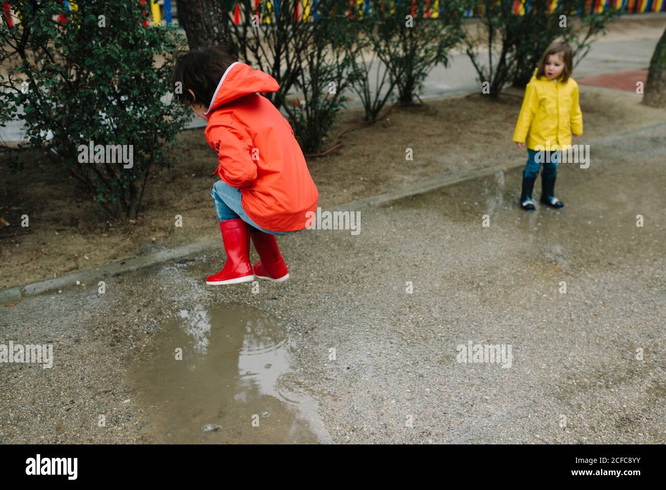 Liebenswert fröhliche Kinder in rot und gelb Regenmantel und Gummi Stiefel mit Spaß spielen in Pfütze in der Straße im Park In grauem Tag Stockfoto
