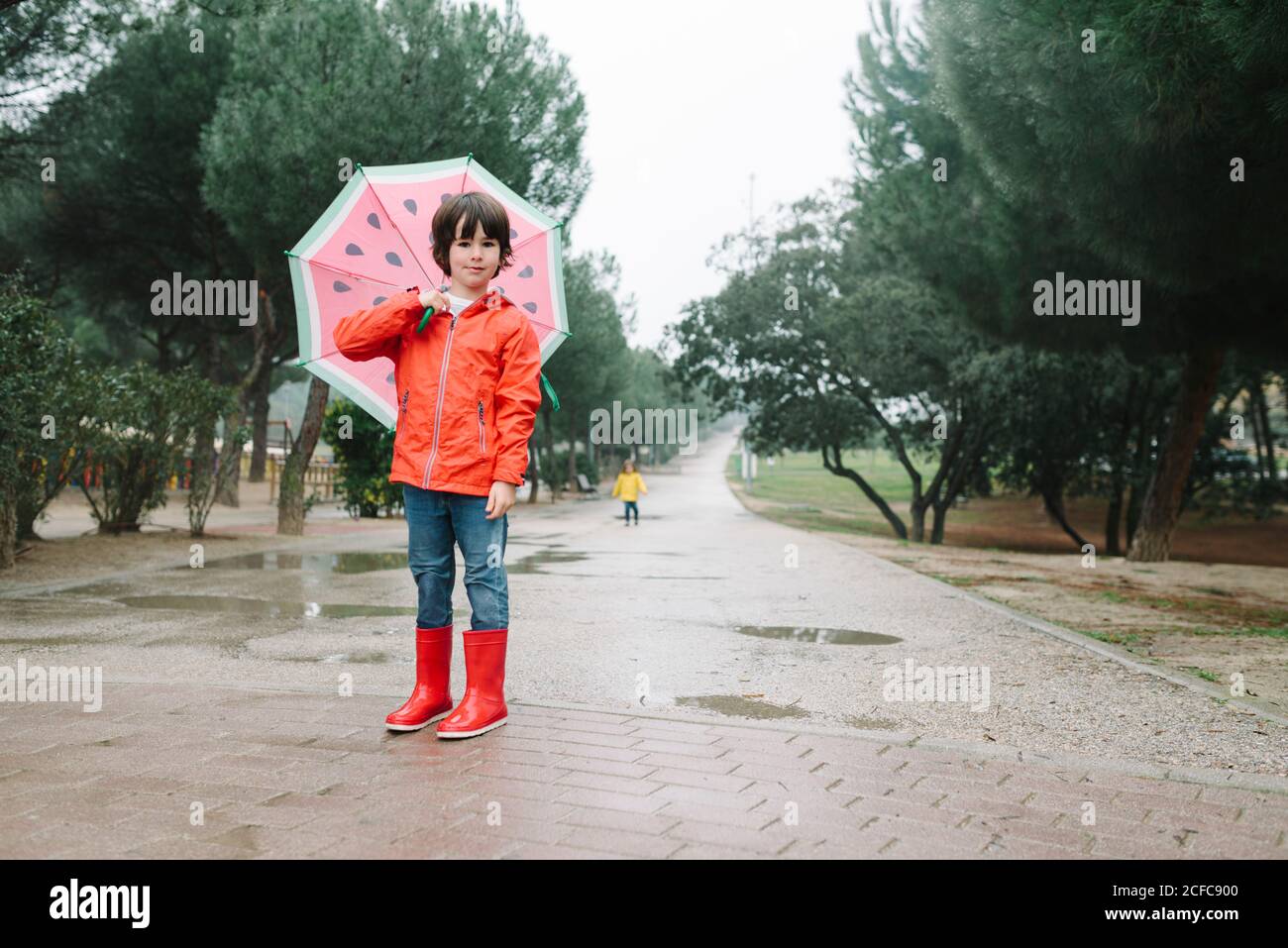 Aktives Kind mit Wassermelone Stile offenen Regenschirm in roten Regenmantel Und Gummistiefel Blick auf Kamera in Parkallee in Grauer Tag Stockfoto